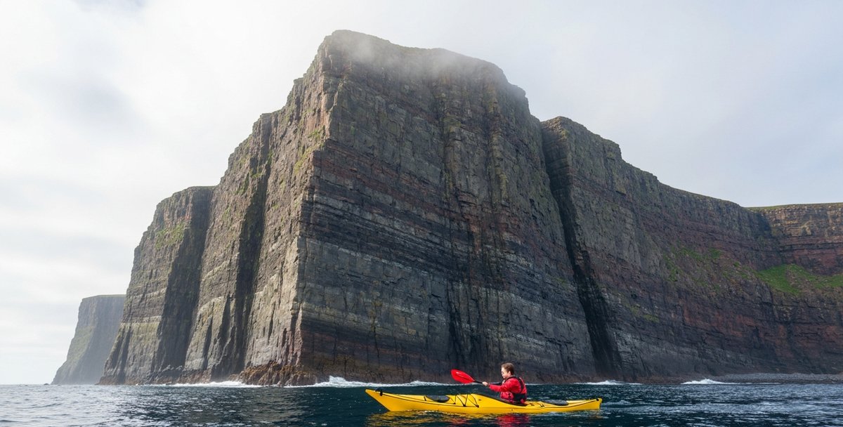 Eye-level sea kayaking photograph below the cliffs at Sliabh Liag on the south-west Donegal coast, Ireland, on a calm summer morning, a solo paddler in a yellow sit-in sea kayak with full spray skirt small in the foreground against the enormous scale of the cliffs rising 601 metres above, dramatic dark sandstone strata visible in the cliff face, mild Atlantic swell, sea-mist clinging to the upper cliff edge, soft Irish morning overcast light, no logos or readable text, paddler photographed from behind with face not visible